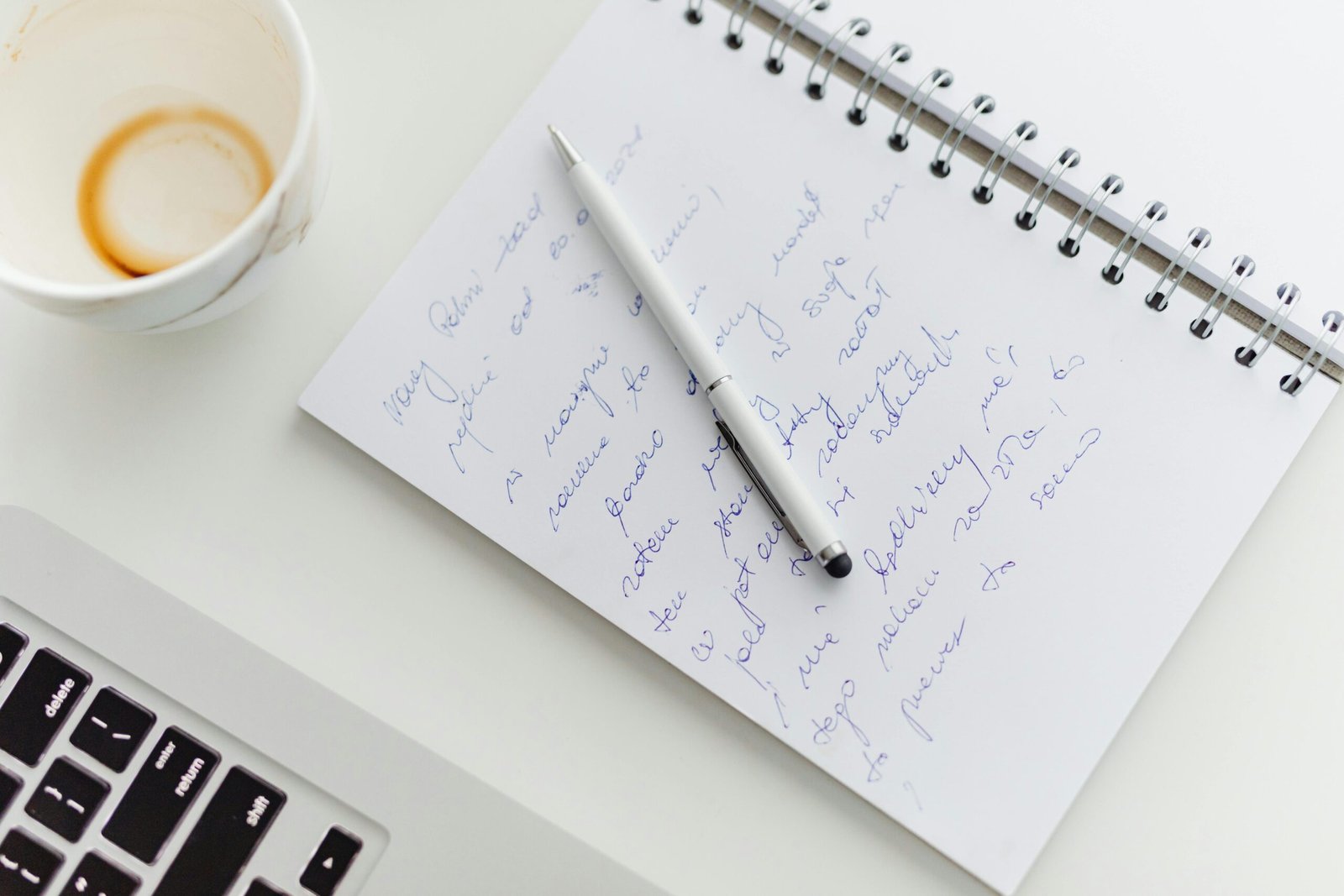 Close up of a notebook with handwriting, a pen, coffee cup, and laptop on a white desk.